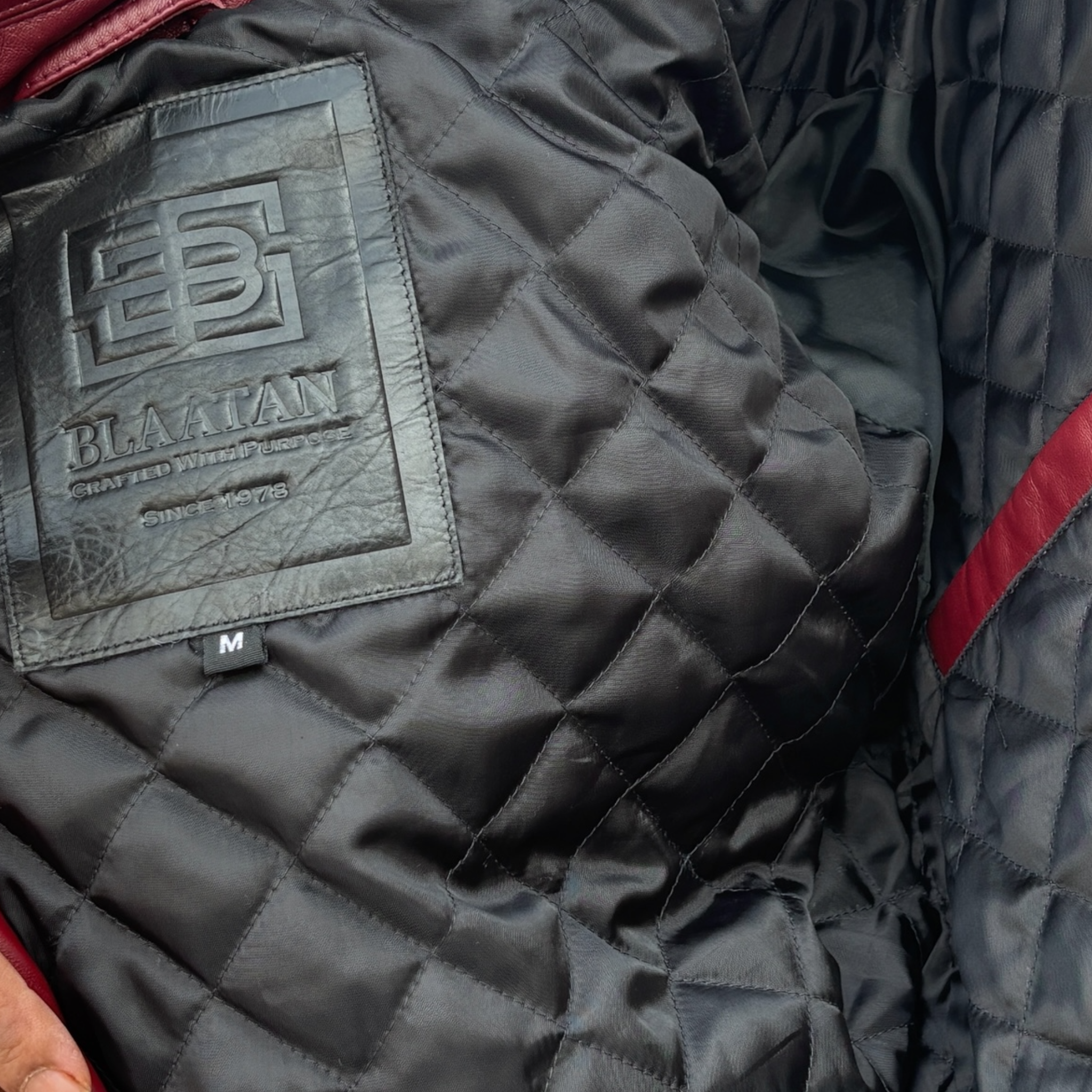 Close-up of a quilted jacket with a brand label on a dark background Blaatan The Garnet Crimson Edition Crimson Burgundy Leather Jacket for Men